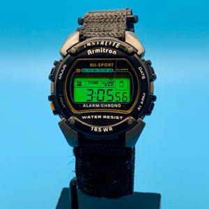 May include: A black Armitron All-Sport digital watch with a green backlit display showing 3:05:56. The watch features a black fabric strap, a round bezel with the text "WATER RESIST 165 WR," and side buttons labeled MODE, DATE, ALARM, and LIGHT.