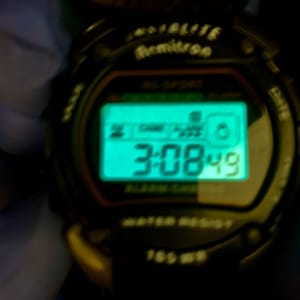 May include: A black digital Armitron Indiglo watch with a glowing teal rectangular screen displaying the time 3:08:49. The watch face features the text Armitron, Indiglo, Alarm Chrono, and Water Resist 165 WR printed in white on the black casing.