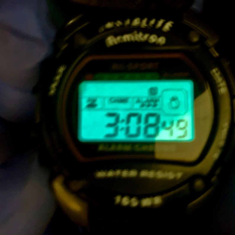 May include: A black digital Armitron Indiglo watch with a glowing teal rectangular screen displaying the time 3:08:49. The watch face features the text Armitron, Indiglo, Alarm Chrono, and Water Resist 165 WR printed in white on the black casing.