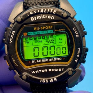 May include: A black Armitron All-Sport digital watch with a bright green backlit display showing 00:00. The watch face features text including Indiglo, Alarm, Chrono, and Water Resist 165 WR. It has a black textured strap and side buttons.
