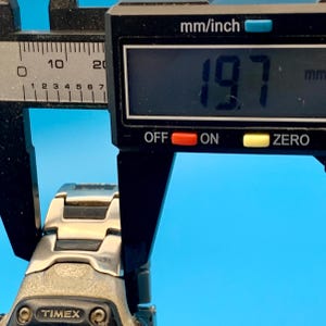 May include: A silver and black metal watch band with a Timex logo, being measured by a black digital caliper showing a reading of 19.7 mm. The watch band features a multi-link design with alternating silver-tone and black-tone segments.
