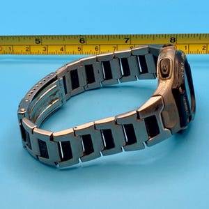 May include: A silver-tone metal watch with a black face and Indiglo feature, positioned next to a yellow measuring tape showing a length of approximately 8.5 inches. The watch band consists of rectangular links with a secure folding clasp.