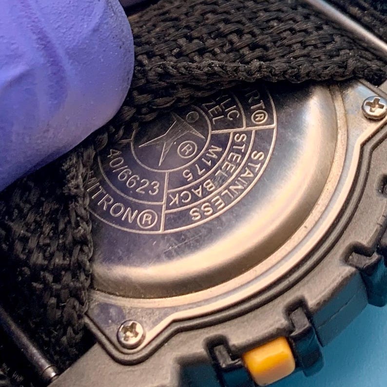 May include: The stainless steel back of a watch case featuring engraved text including "STAINLESS STEEL BACK", "M175", "40/6623", and "WITRON". A black woven strap is attached to the watch, and a small yellow button is visible on the side of the case.