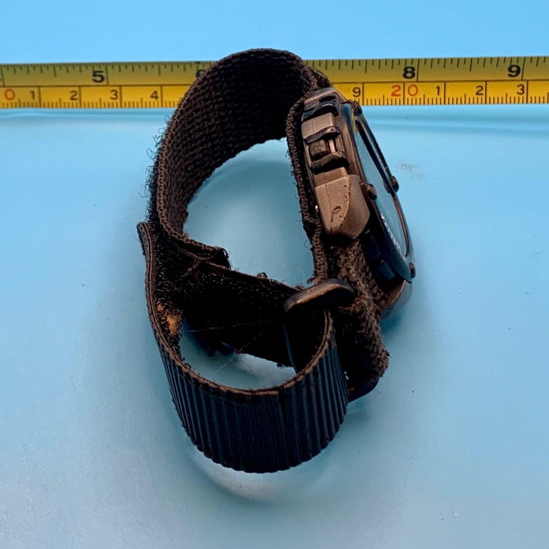 May include: A black digital watch with a textured fabric strap and a hook-and-loop closure. A yellow measuring tape is positioned behind the watch, showing the watch case width is approximately 1.5 inches and the strap length is 9 inches.