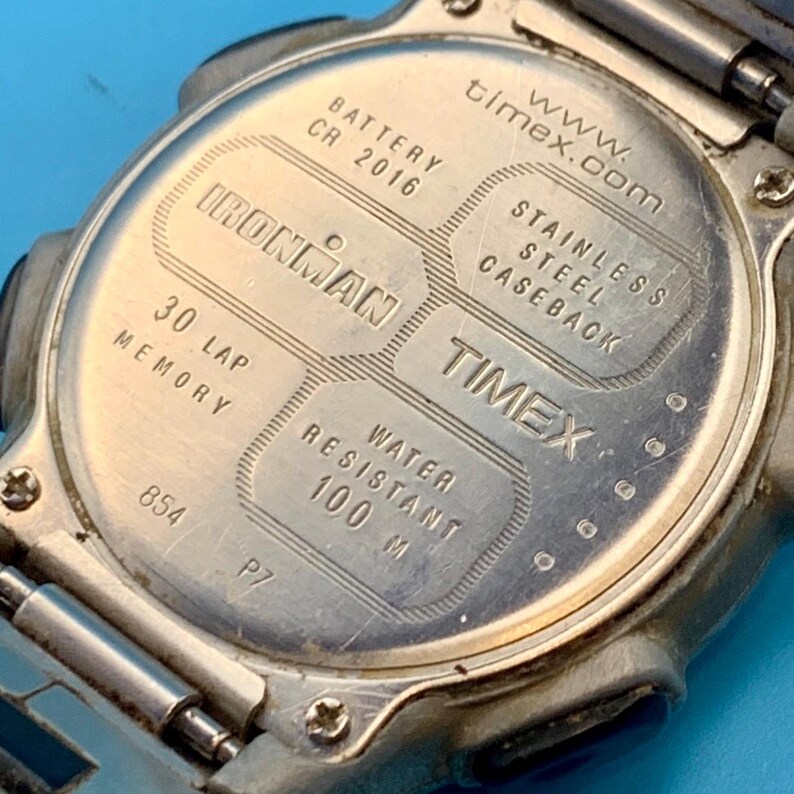 May include: The silver-toned stainless steel caseback of a Timex Ironman watch. Engraved text includes Ironman, 30 Lap Memory, Water Resistant 100 M, Battery CR 2016, and www.timex.com. Four small screws secure the circular metal back plate.