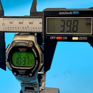 May include: A silver and black Timex Ironman 30 Lap digital watch with a green backlit display showing the time 10:37 and date SU 1/07. A black digital caliper is positioned over the watch, displaying a measurement of 39.8 mm on its screen.