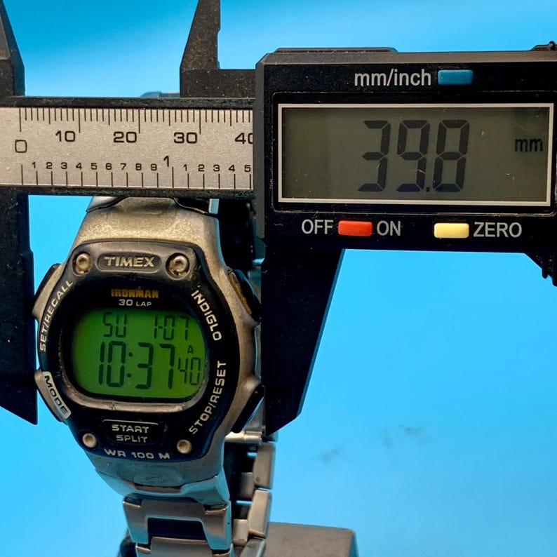 May include: A silver and black Timex Ironman 30 Lap digital watch with a green backlit display showing the time 10:37 and date SU 1/07. A black digital caliper is positioned over the watch, displaying a measurement of 39.8 mm on its screen.