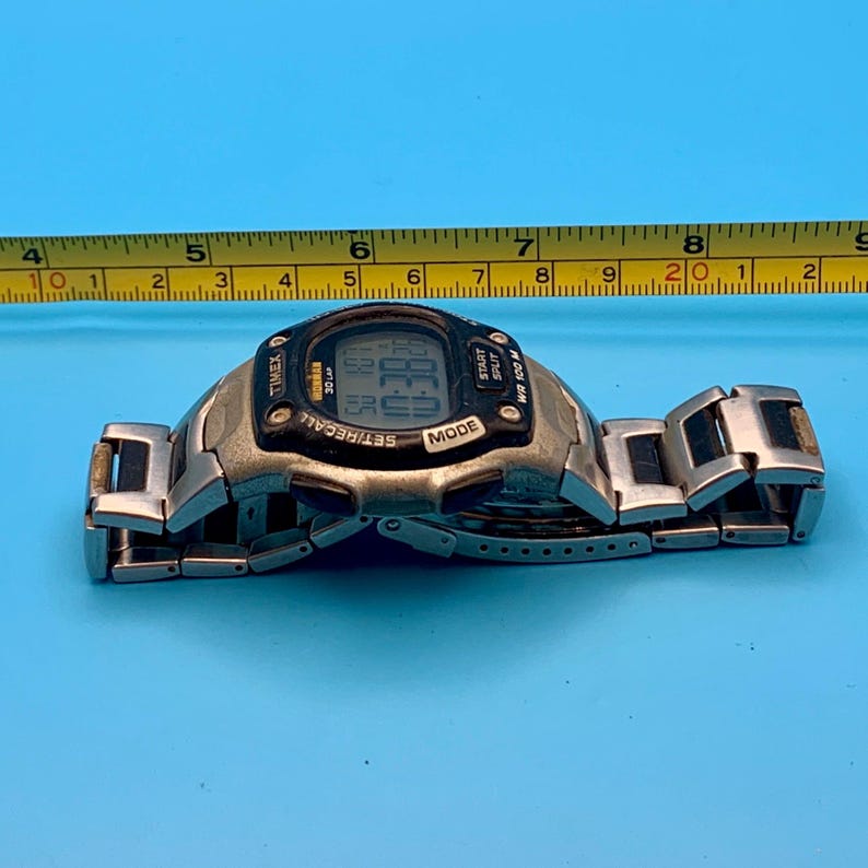 May include: A silver-tone digital wristwatch with a black bezel featuring the text Timex, Ironman, 30 Lap, Start, Split, Mode, and WR 100M. A yellow measuring tape positioned above the watch shows a length of approximately 8.5 inches.
