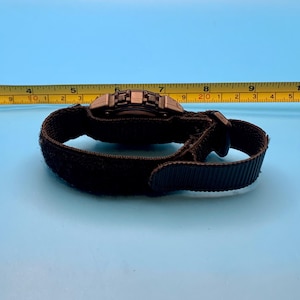 May include: A black digital watch with a textured nylon strap and a hook-and-loop closure. The watch is positioned in front of a yellow measuring tape, showing the watch case width is approximately 1.75 inches across the center of the dial.