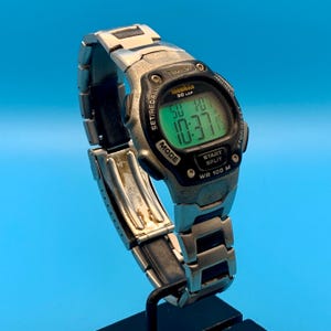 May include: A silver-tone Timex Ironman 30 Lap digital watch with a stainless steel link bracelet. The watch face features a green backlit display showing the time 10:37, the date, and text including Indiglo, Set/Recall, Mode, and Start Split.