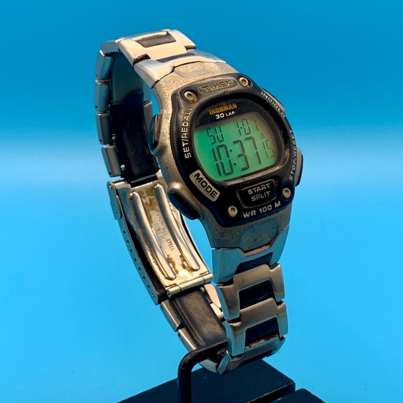 May include: A silver-tone Timex Ironman 30 Lap digital watch with a stainless steel link bracelet. The watch face features a green backlit display showing the time 10:37, the date, and text including Indiglo, Set/Recall, Mode, and Start Split.