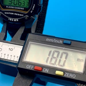 May include: A black digital wristwatch with a green display showing the text ALARM/CHRONO and WATER RESIST 165 WR. The watch is being measured by a black digital caliper displaying a measurement of 18.0 mm on its screen for precise sizing.