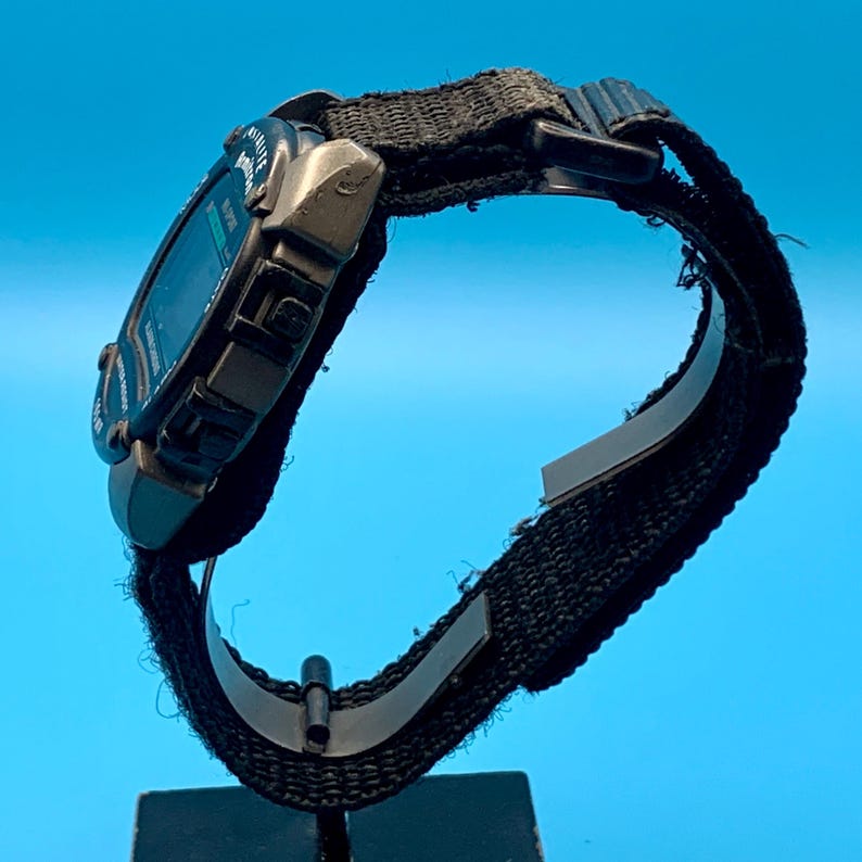 May include: A digital wristwatch with a dark gray rectangular case and a black nylon fabric strap. The watch face features a dark display with visible text including the word Illuminator. The strap is secured with a black plastic buckle.