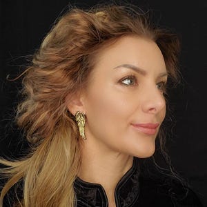 Puede incluir: Una mujer con el pelo largo, ondulado y castaño claro lleva un par de pendientes de alas de ángel doradas. Lleva una chaqueta de terciopelo negro.