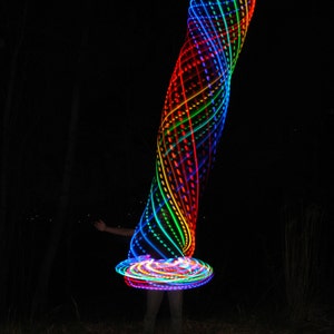 May include: A person is spinning a rainbow colored LED hoop in the dark. The hoop is creating a spiral pattern of light.