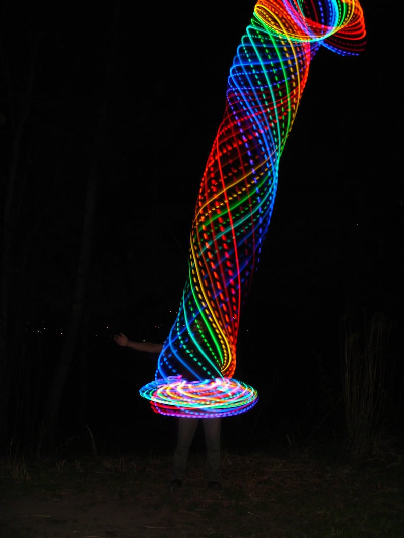 May include: A person is spinning a rainbow colored LED hoop in the dark. The hoop is creating a spiral pattern of light.