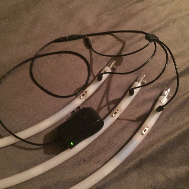 May include: Three white flexible LED light strips with black wires and a black power adapter. The light strips have gold connectors and are connected to a black Y-shaped connector.