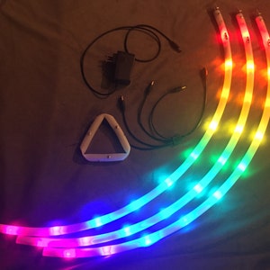 May include: Four flexible LED light strips with different colored lights, including red, orange, yellow, green, blue, and purple. The lights are connected to a power adapter and a triangular controller.