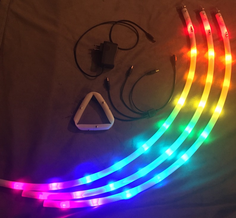 May include: Four flexible LED light strips with different colored lights, including red, orange, yellow, green, blue, and purple. The lights are connected to a power adapter and a triangular controller.