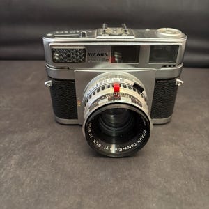 May include: A vintage silver Braun Paxette camera with a black leather grip. The lens reads "BRAUN-Color-Ennalyt SLK 1:1.9/50". The camera has a silver body with the text "PAUK Paselle" on the top.