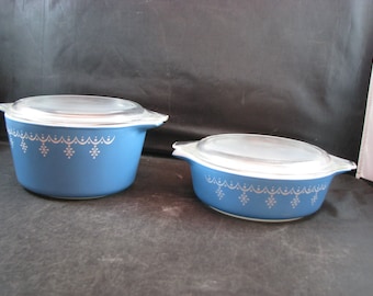 Op de afbeelding: Twee blauwe ovenschaaltjes met witte sneeuwvlokmotieven en heldere glazen deksels. De grotere schaal is op de voorgrond en de kleinere is op de achtergrond.