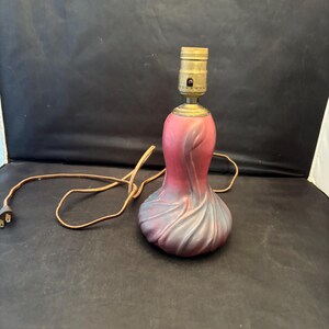 May include: A small, decorative table lamp with a pink and purple base and a brass socket. The base has a textured, organic design. The lamp has a brown electrical cord with a two-prong plug.