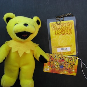 May include: Yellow plush bear with a black mouth and eyes. The bear is wearing a yellow collar with a star design. The bear is holding a yellow tag with the text "Wharf Rat" and a red heart. The tag is attached to a chain with a small metal tag that reads "Limited Edition 06211 / 25000 Grateful Dead 2000 Tour Admission".