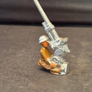 May include: A silver toy soldier figurine holding a silver toy anti-aircraft gun. The soldier is wearing a brown uniform and a helmet. The gun has the letters "U.S." on the side.