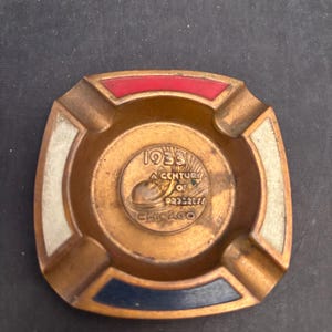 May include: Vintage bronze ashtray with a square shape and rounded corners. The ashtray features red, white, and blue accents on the corners. The center has the text "1933 A Century of Progress Chicago".