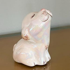 May include: A ceramic bunny figurine in a seated position, with its head tilted upwards. The bunny is primarily white with iridescent highlights, and features pink accents on the nose and ears. Brown details define the eyes and nose.