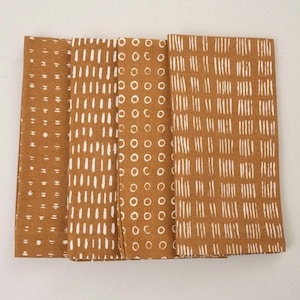 May include: Four folded brown cloth napkins with white patterns. Each napkin features a different design: small dashes, vertical lines, circles, and a grid of vertical lines. The napkins are arranged side by side.