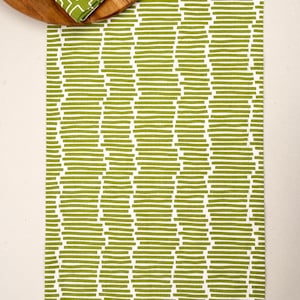 May include: A rectangular table runner featuring a repeating pattern of horizontal olive green bars on a white background. A round wooden tray sits at the top, holding folded napkins with matching green and white geometric and floral prints.