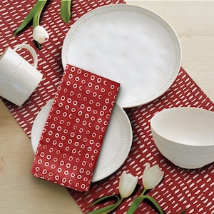 May include: A white ceramic mug, plates, and bowl are arranged on a red patterned table runner. A red napkin with white circles is placed on a plate. White tulips add a touch of freshness to the dining setting. The tableware has a dotted rim.