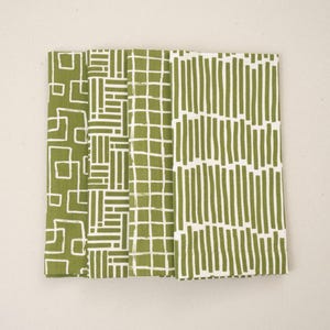 May include: Four folded green and white patterned cloth napkins. The napkins feature different geometric designs, including squares, rectangles, and vertical lines. The color scheme is a light olive green and white.