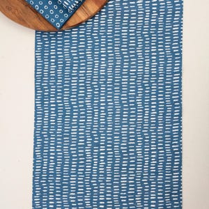 May include: A rectangular blue table runner featuring a repeating pattern of short, white, hand-painted style dashes. A folded blue napkin with a matching dash pattern rests on a circular wooden board positioned at the top left of the runner.