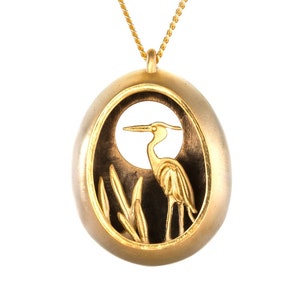 May include: Gold pendant necklace featuring a detailed cutout of a heron standing in reeds. The pendant is oval shaped and has a dark background.