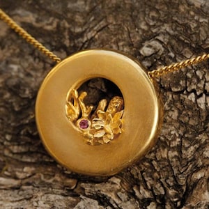 以下が含まれることがあります： 小さなピンクの宝石が中央に飾られた、繊細な花のデザインが施された丸いディスクが特徴のゴールドのペンダントネックレス。ペンダントはゴールドチェーンから吊り下げられています。