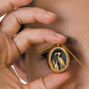 May include: A gold oval pendant necklace with a detailed image of a heron standing in reeds. The pendant is held in a person's hand.