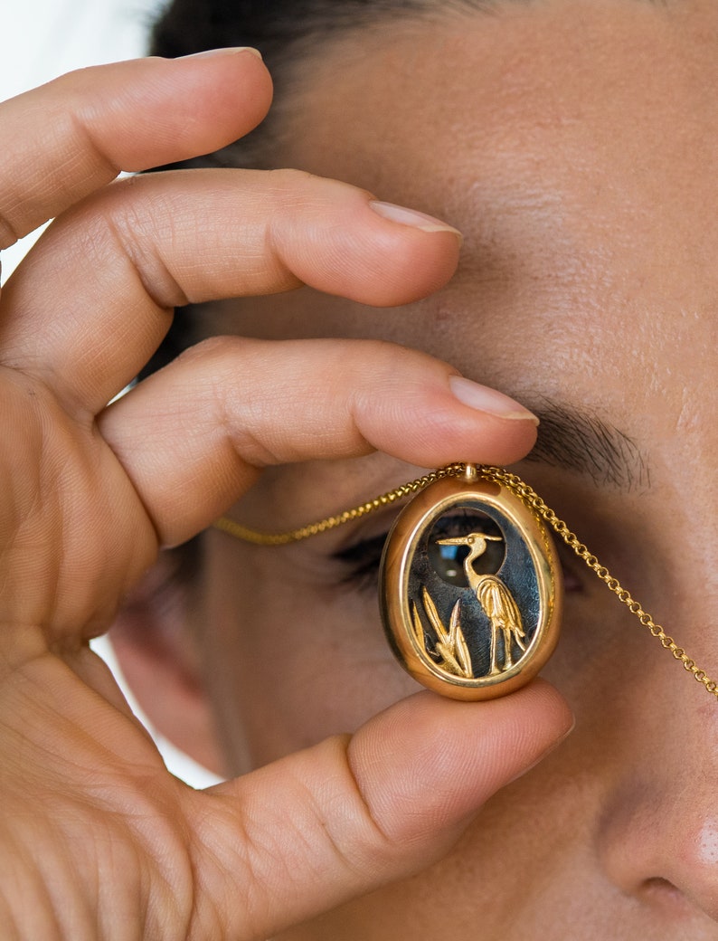 May include: A gold oval pendant necklace with a detailed image of a heron standing in reeds. The pendant is held in a person's hand.