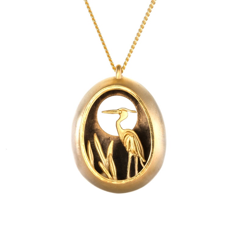 May include: Gold pendant necklace featuring a detailed cutout of a heron standing in reeds. The pendant is oval shaped and has a dark background.