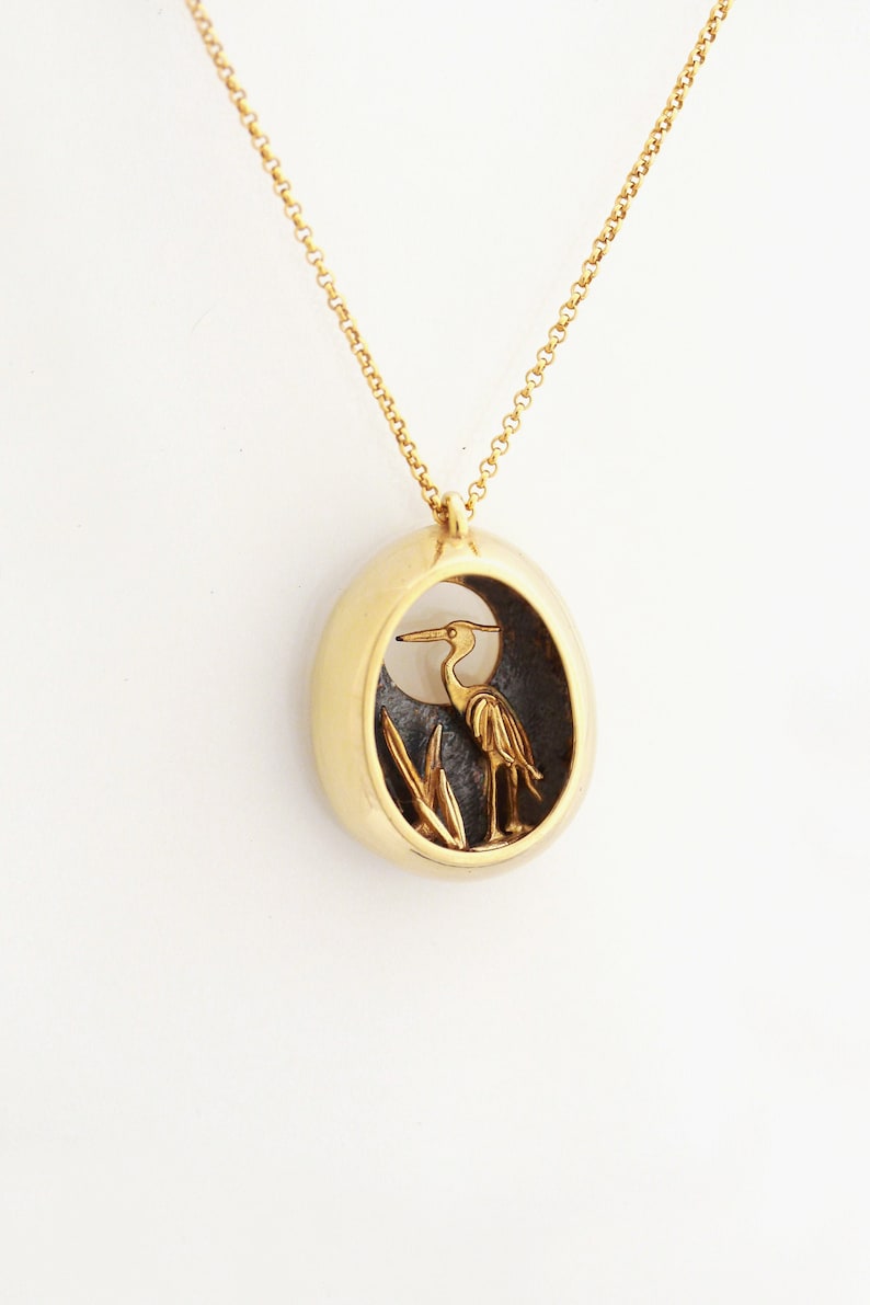 May include: A gold necklace with a pendant featuring a detailed silhouette of a heron standing in reeds against a black background.