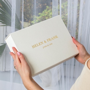 以下が含まれることがあります： クリーム色の長方形の箱で、ヒンジ付きの蓋が付いています。「HELEN & FRANK」と「JUNE 20, 2026」と金色の文字で書かれています。箱は、白いシアーなカーテンのある窓の前で、2つの手で持たれています。