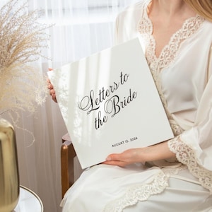 以下が含まれることがあります： 「Letters to the Bride」と書かれた白い本。日付は2026年8月15日。白いレースのローブを着た人が本を持っています。背景には窓とドライフラワーがあります。