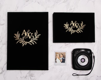 May include: Two black photo albums with gold lettering and a floral design. The albums say "A&V" on the front. A white photo frame with a photo of a couple in it and a black instant camera are also in the image.