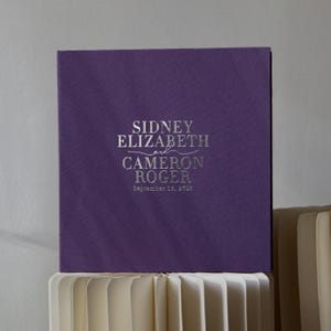 May include: A square, deep purple wedding album with silver lettering. The text reads "SIDNEY ELIZABETH and CAMERON ROGER" with the date "September 18, 2023" below. The album is resting on a stack of cream-colored paper.