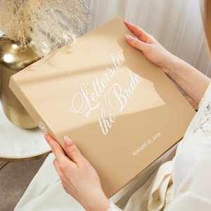 Pode incluir: Uma caixa bege com as palavras "Letters to the Bride" em escrita branca elegante. A data "August 15, 2026" está impressa abaixo. A caixa é segurada por uma pessoa vestindo um roupão branco.