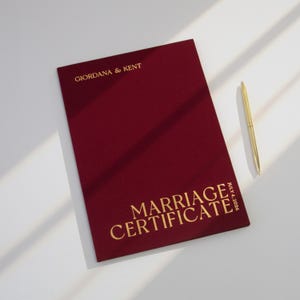 May include: A deep red, rectangular marriage certificate folder with gold foil lettering that reads GIORDANA & KENT and MARRIAGE CERTIFICATE JULY 4, 2026. A slim gold pen is positioned to the right of the folder on a plain, light-colored surface.