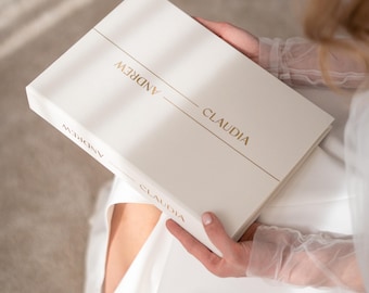 May include: A rectangular white book with a smooth matte finish. The front cover features the names ANDREW and CLAUDIA printed in gold foil lettering, separated by a thin horizontal gold line. The spine also displays the names in gold.