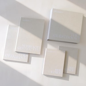 Può includere: Un set di cinque libri beige chiaro con testo bianco in rilievo. Le copertine sono etichettate come The Guestbook, Marriage Certificate, The Officiant, Her Vows e His Vows. Ogni libro presenta il testo The Linney's in alto in un carattere sans-serif.