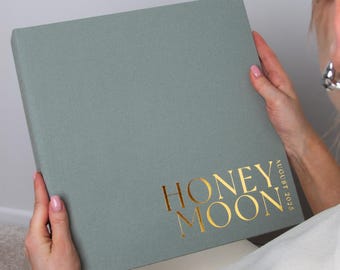 Könnte beinhalten: Ein quadratisches Fotoalbum in Salbeigrün mit matter Oberfläche. Auf dem Vorderdeckel befindet sich der in Goldfolie geprägte Schriftzug "HONEY MOON" in einer fetten Serifenschrift, ergänzt durch den vertikalen Golddruck "AUGUST 2025" rechts.